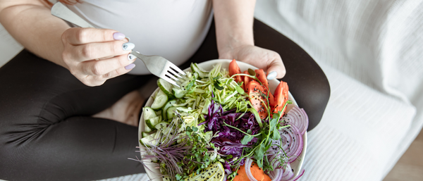 A pregnant woman in leggings and a white top sits with a colorful salad bowl, holding a fork.