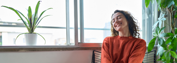 A woman in an orange sweater sits in a bright room near a window, smiling with her eyes closed as she basks in the natural sunlight.