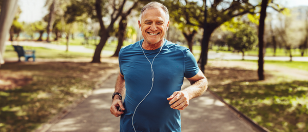 A senior man with a fit physique, wearing a blue athletic shirt and earphones, jogging on a paved path in a green park while smiling.