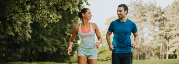 A smiling man and woman jogging together in a lush green park, wearing athletic clothing.