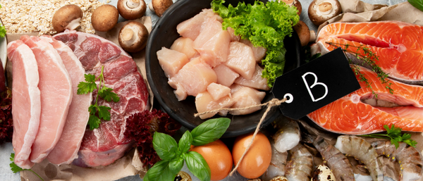 A variety of vitamin B-rich foods including raw salmon steaks, chicken breast pieces in a black bowl, pork and beef cuts, shrimp, eggs, mushrooms, oats, and fresh herbs such as basil and parsley, arranged on a rustic surface with a black tag labeled