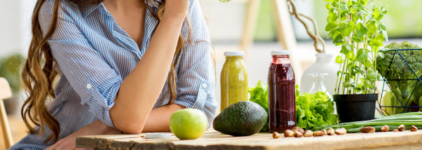 A woman in a blue striped shirt sits at a wooden table surrounded by fresh fruits, vegetables, green herbs, and two colorful juice bottles. 