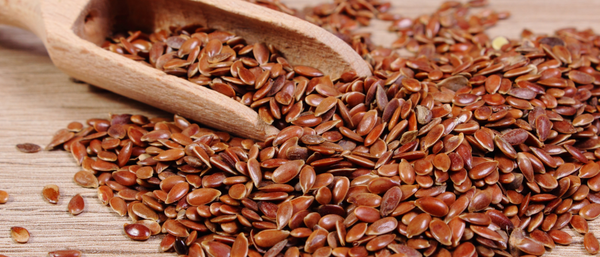 A close-up of glossy brown flax seeds spilling from a rustic wooden scoop onto a wooden surface. 