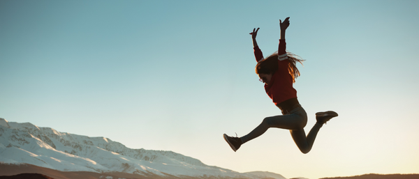 A young woman jumps energetically in the air against a stunning backdrop of snow-capped mountains during sunset, symbolizing freedom, adventure, and happiness.